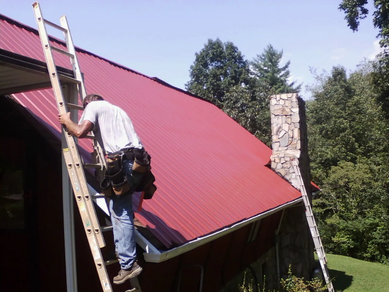 Roofer on a ladder beside a red metal roof and stone chimney — Soffit & Fascia Repair by Roofing Brothers in Munfordville